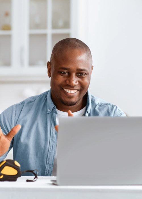 Happy african american handyman recording video about repairs and construction tools, looking at laptop webcam and sitting in kitchen interior with equipment on table, empty space